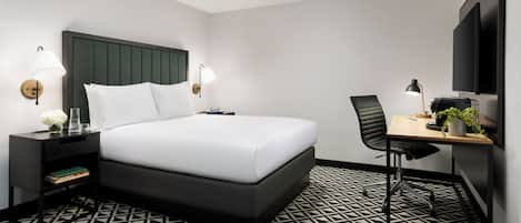 A modern hotel room with a large bed, a desk with a chair, a flat-screen TV, and a patterned floor.