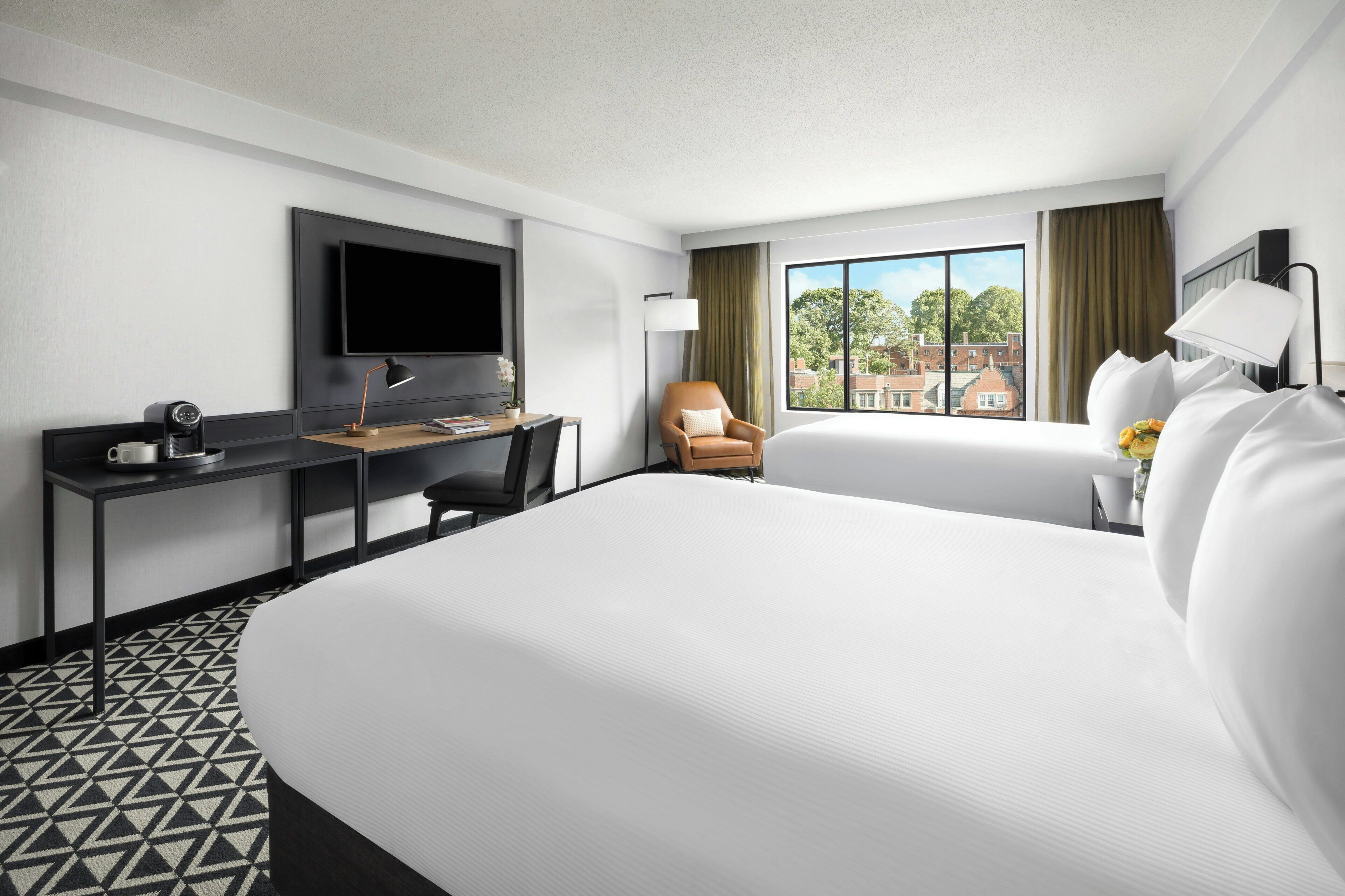 A hotel room with a large bed, a desk with a flat-screen TV, a chair, and a window with a view of buildings.