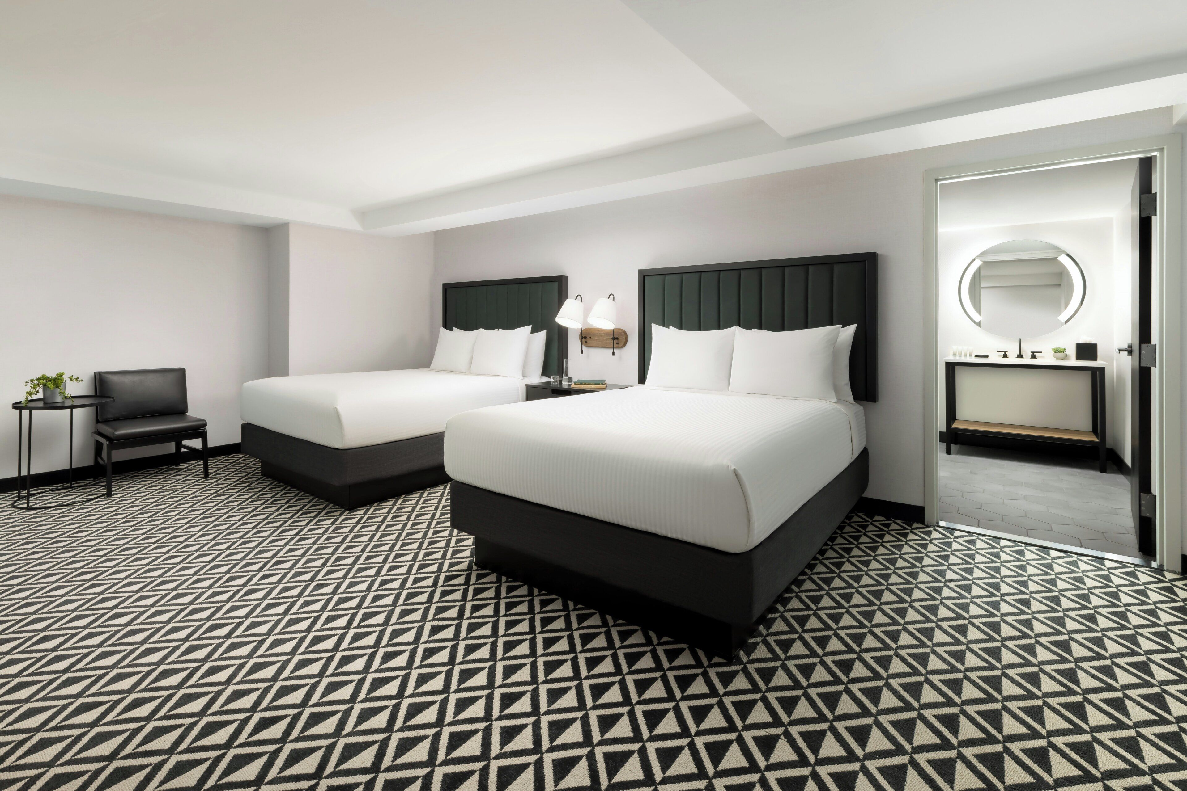 A hotel room with two beds, a small table with a chair, a bedside lamp, and a bathroom with a mirror.