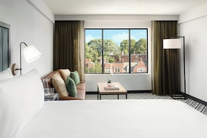 A hotel room with a large bed, a small table with a book, a lamp, and a view of buildings through a window. - Arcadian Hotel (Brookline)