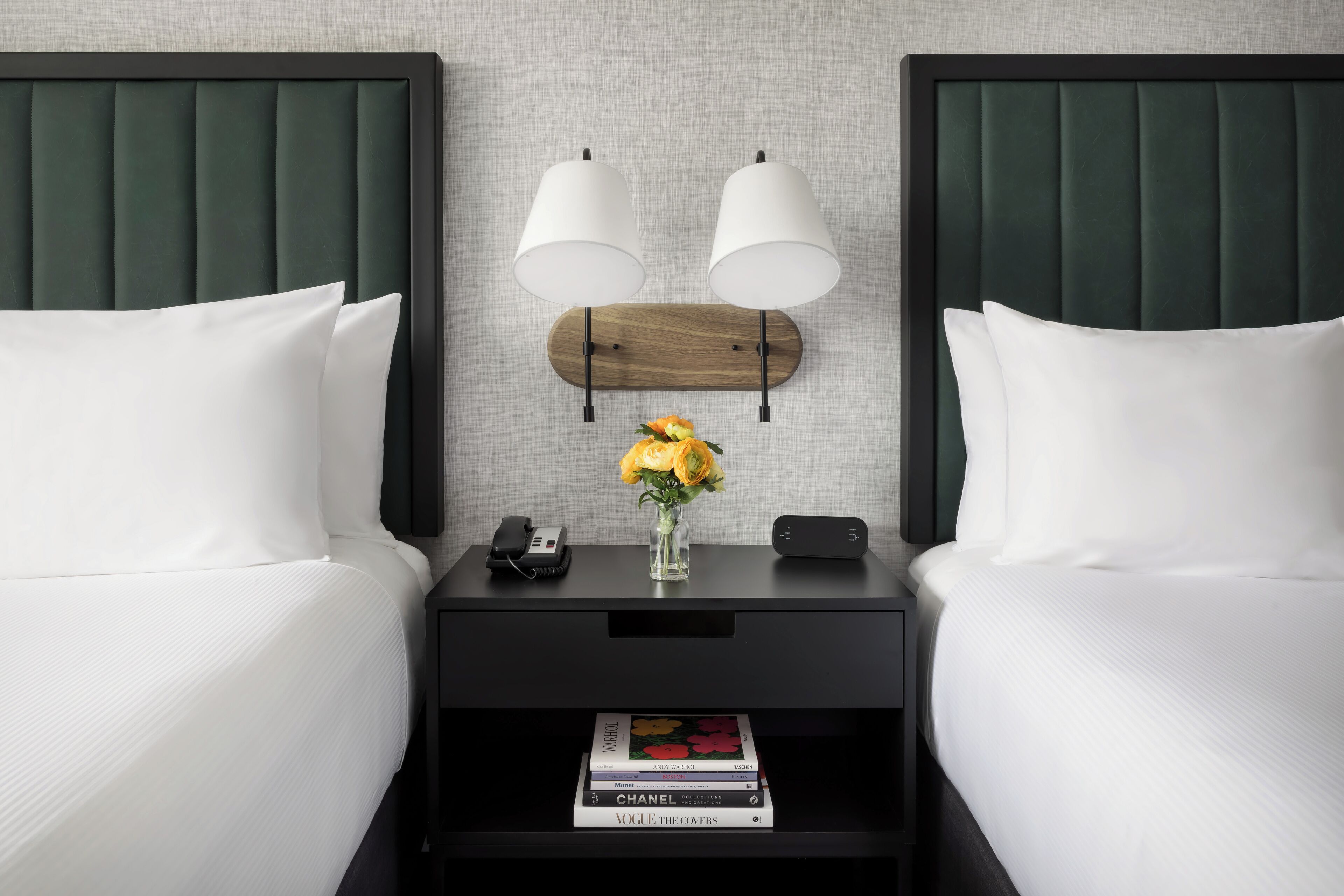 A hotel room with two beds, a nightstand with a phone and a vase of flowers, and a bookshelf with magazines.