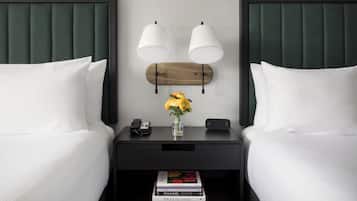 A hotel room with two beds, a nightstand with a phone and a vase of flowers, and a bookshelf with magazines.