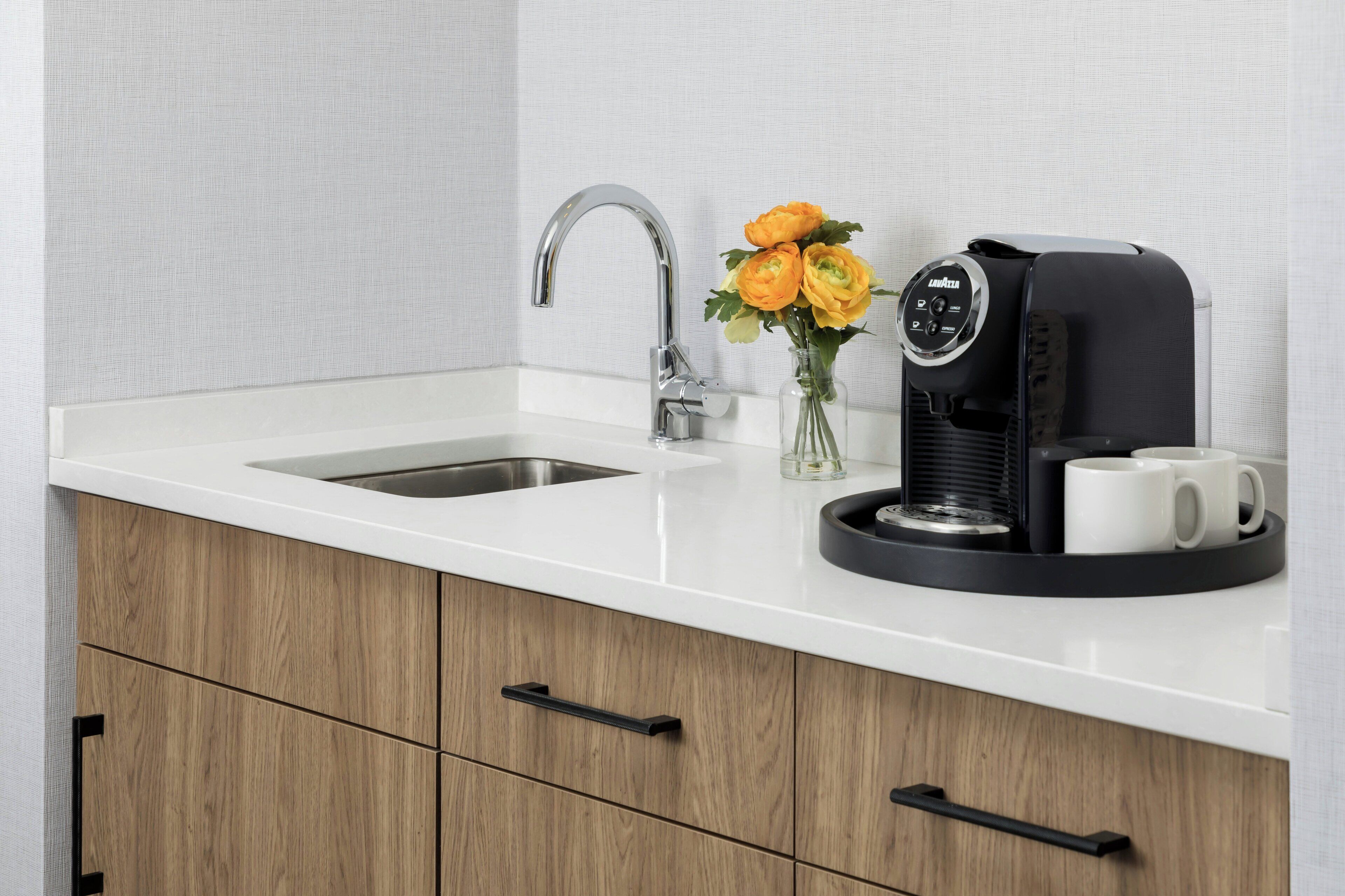 A kitchen with a white countertop, a stainless steel sink, a coffee machine, and a vase with flowers.