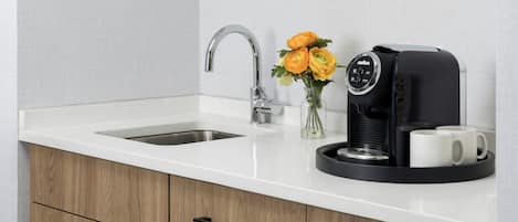 Uma cozinha com bancada branca, pia de aço inoxidável, cafeteira e um vaso com flores.