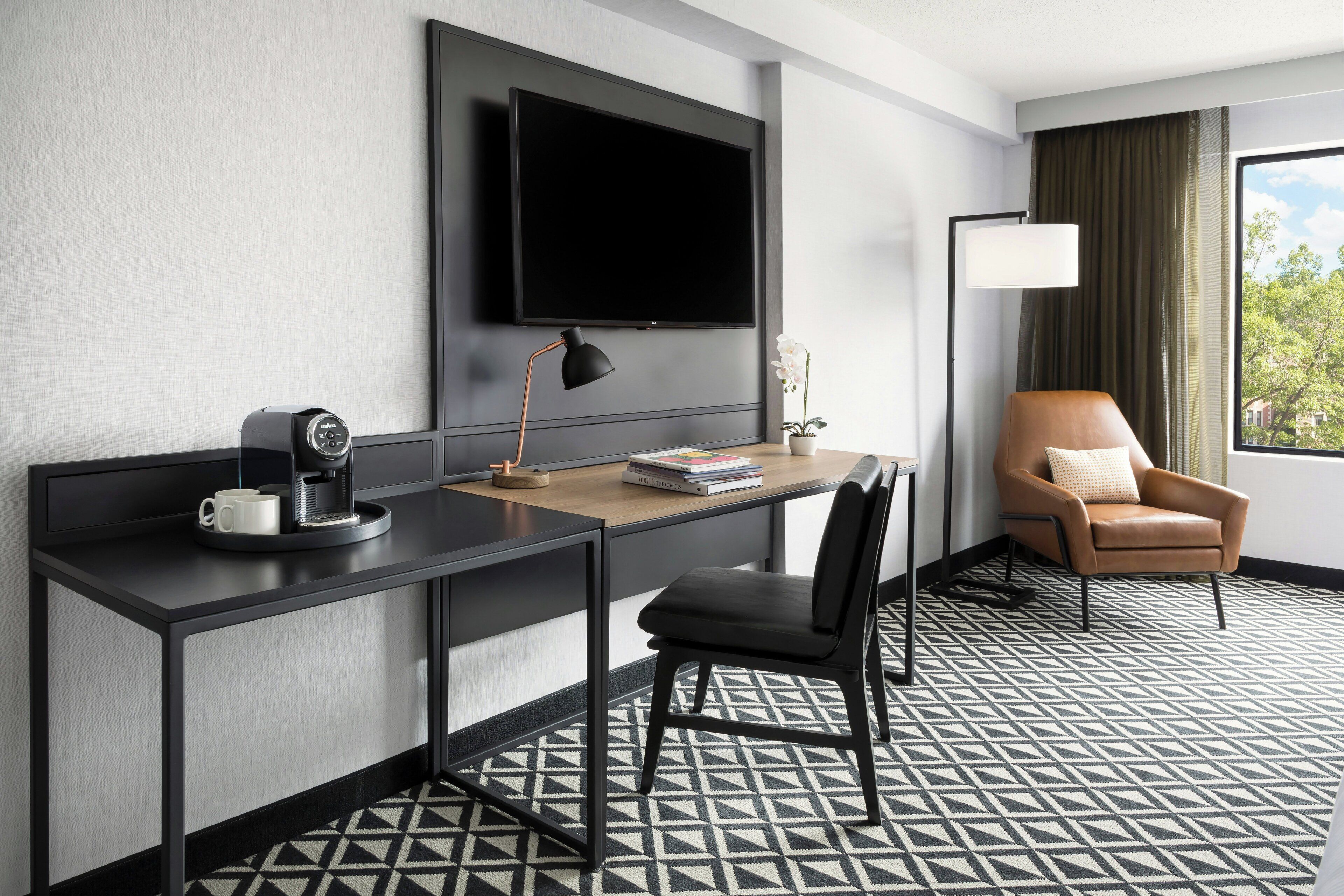 A hotel room with a desk, a chair, a television mounted on the wall, a lamp, and a window with curtains.