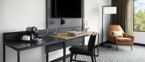 Une chambre d’hôtel équipée d’un bureau, d’une chaise, d’un téléviseur fixé au mur, d’une lampe et d’une fenêtre avec des rideaux.