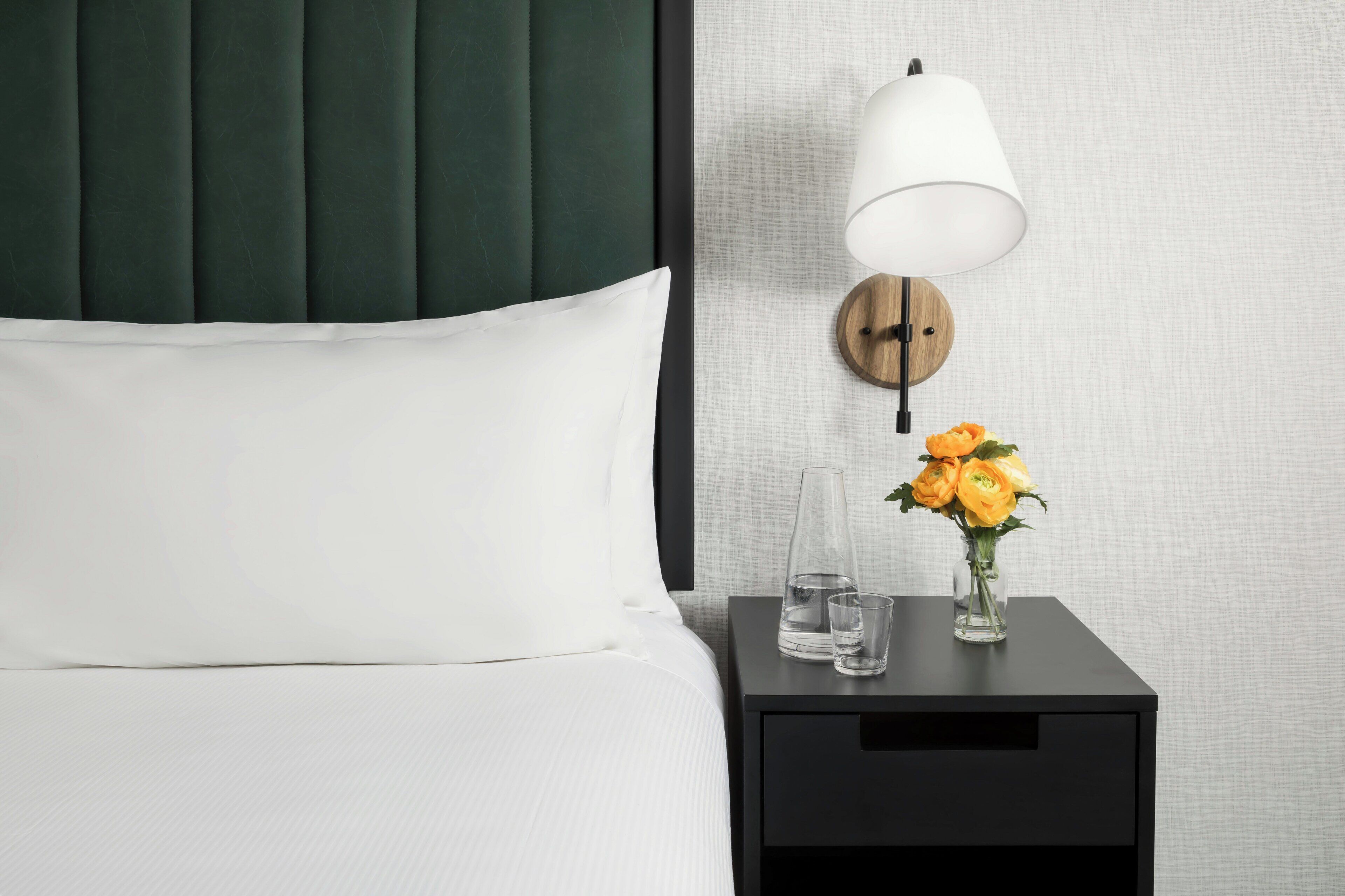 Een netjes opgemaakt bed met witte lakens en kussens, een nachtkastje met een bloemenvaas, een glas water en een wandlamp.