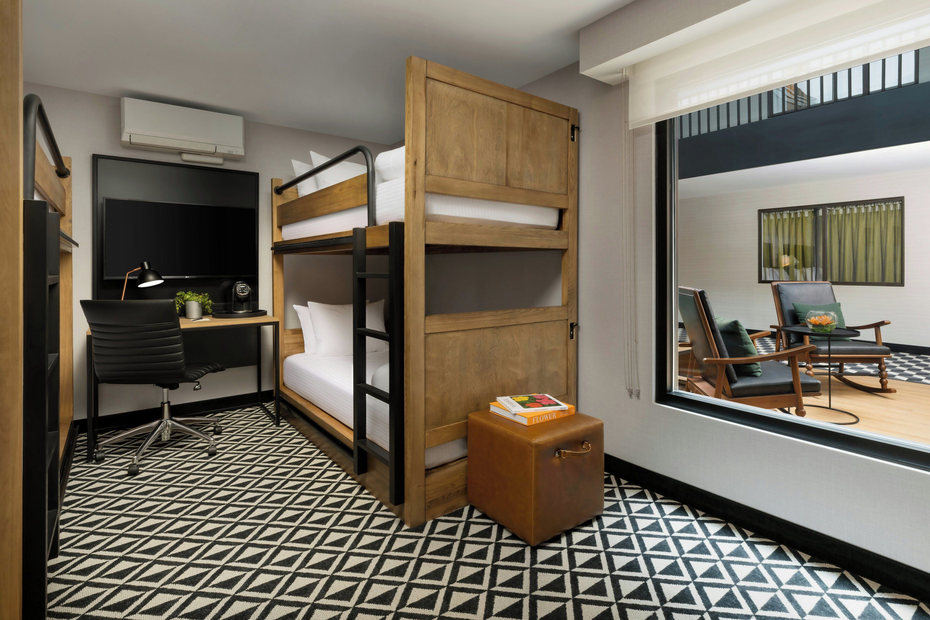 A compact hotel room with a bunk bed, a desk with a chair, a small table with a plant, and a suitcase.