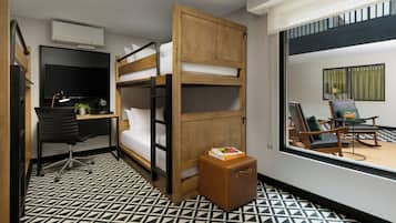 A compact hotel room with a bunk bed, a desk with a chair, a small table with a plant, and a suitcase.