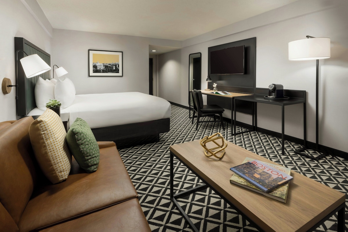 A hotel room with a bed, a desk, a chair, a coffee table, a TV, and a lamp.
