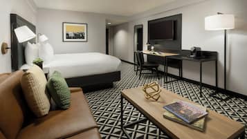 A hotel room with a bed, a desk, a chair, a coffee table, a TV, and a lamp.