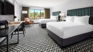 A hotel room with two beds, a desk, a chair, a TV, and a large window.