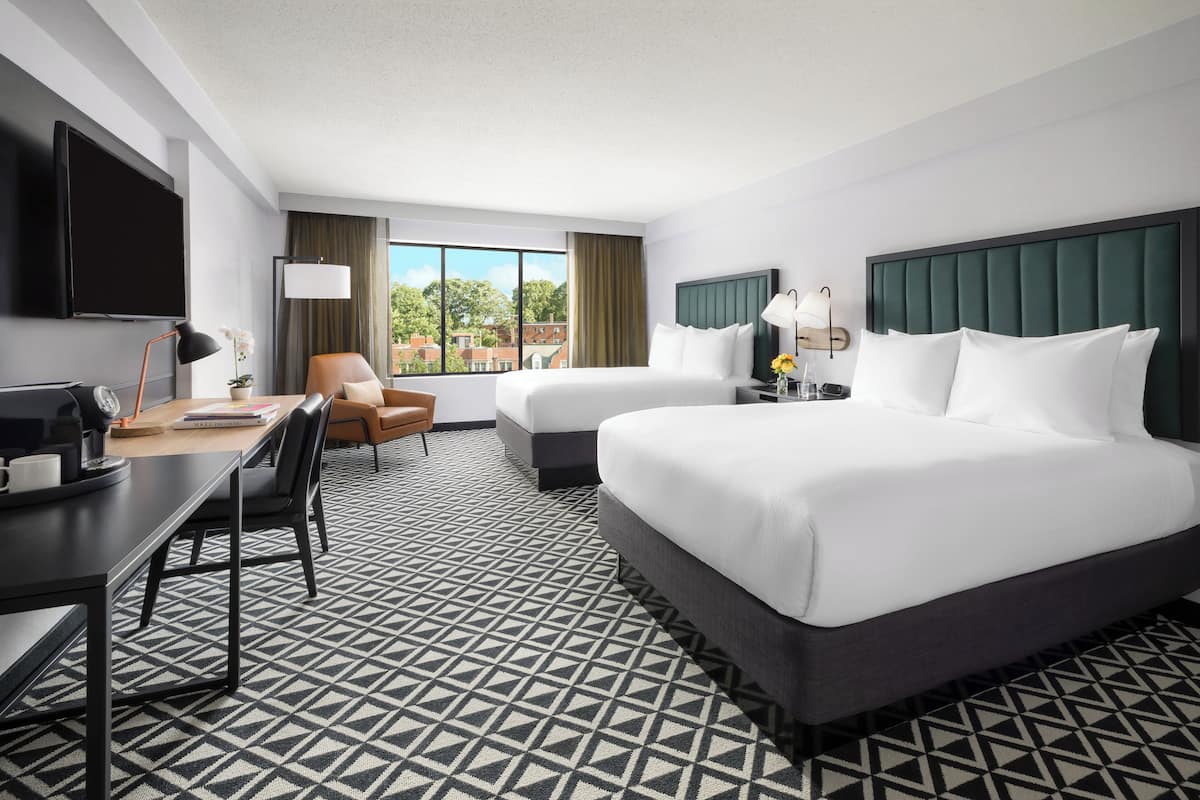 A hotel room with two beds, a desk, a chair, a TV, and a large window.