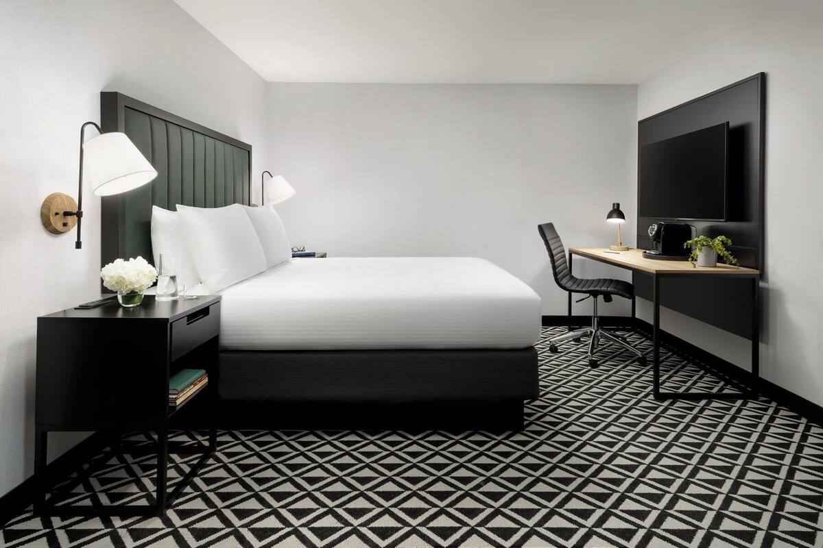 A modern hotel room with a bed, two bedside lamps, a desk with a chair, a TV, and a patterned floor.