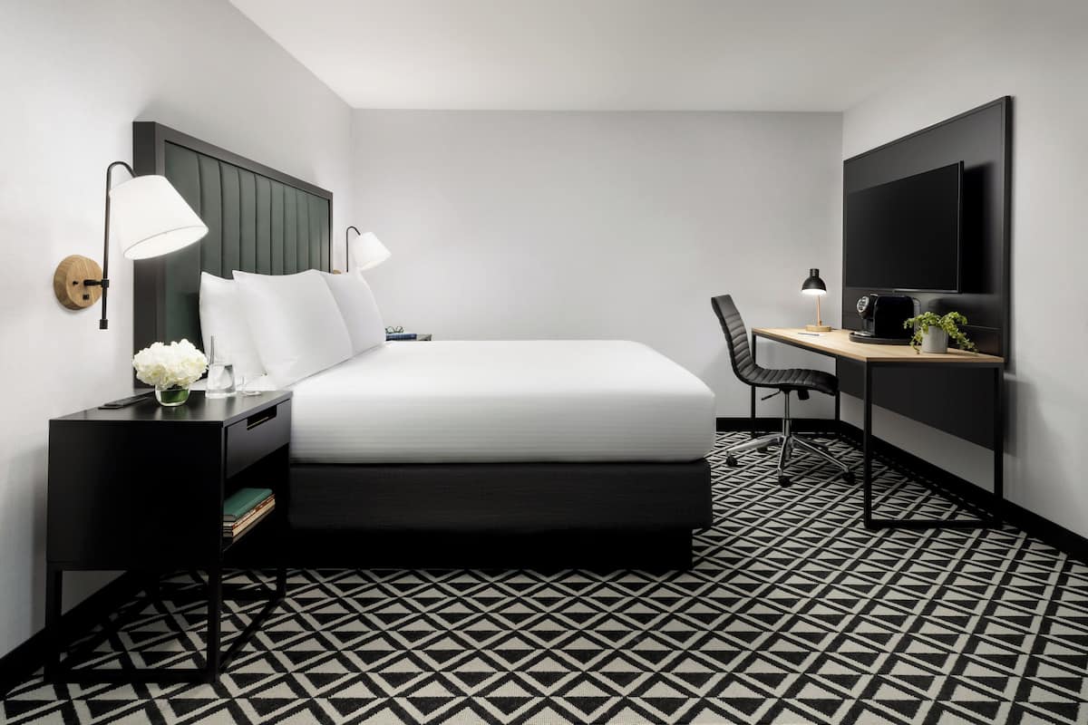A modern hotel room with a bed, bedside tables, a desk, a chair, a TV, and a patterned floor.