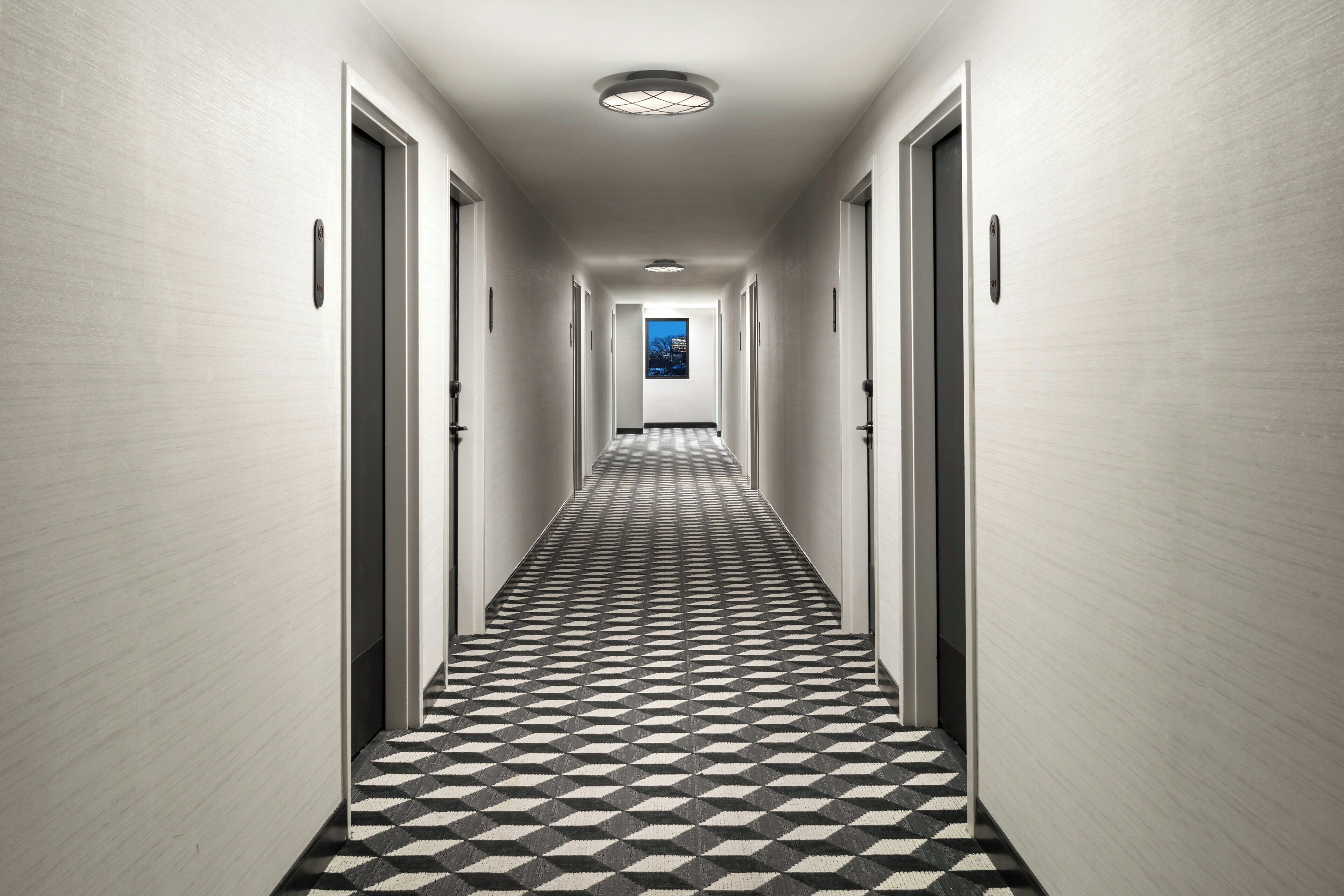 A hallway with a checkered floor pattern, doors on both sides, and a view of the outside at the end.