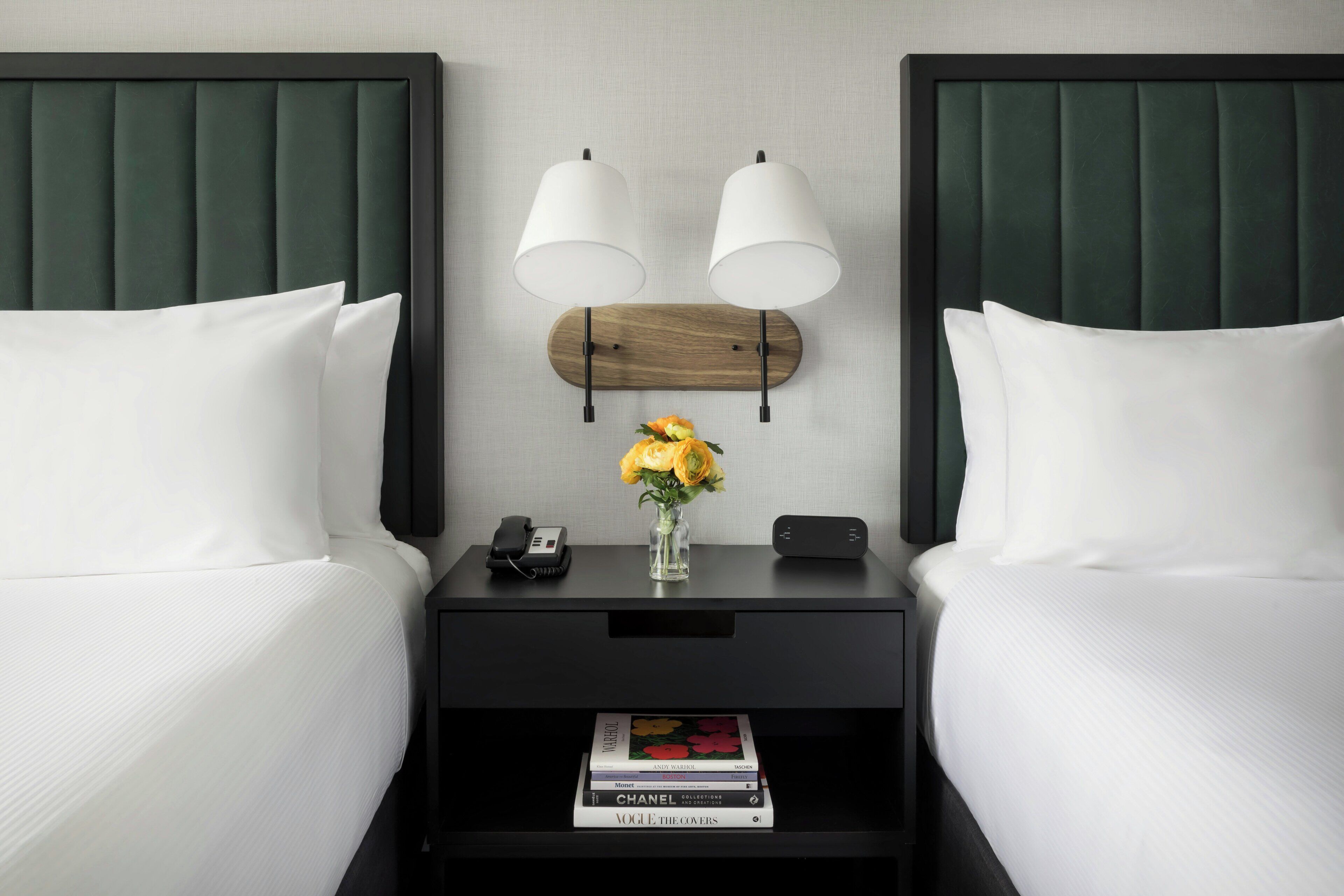 Une chambre d’hôtel avec deux lits, une table de chevet avec une lampe, un téléphone, un vase avec des fleurs et un livre sur la table de chevet.
