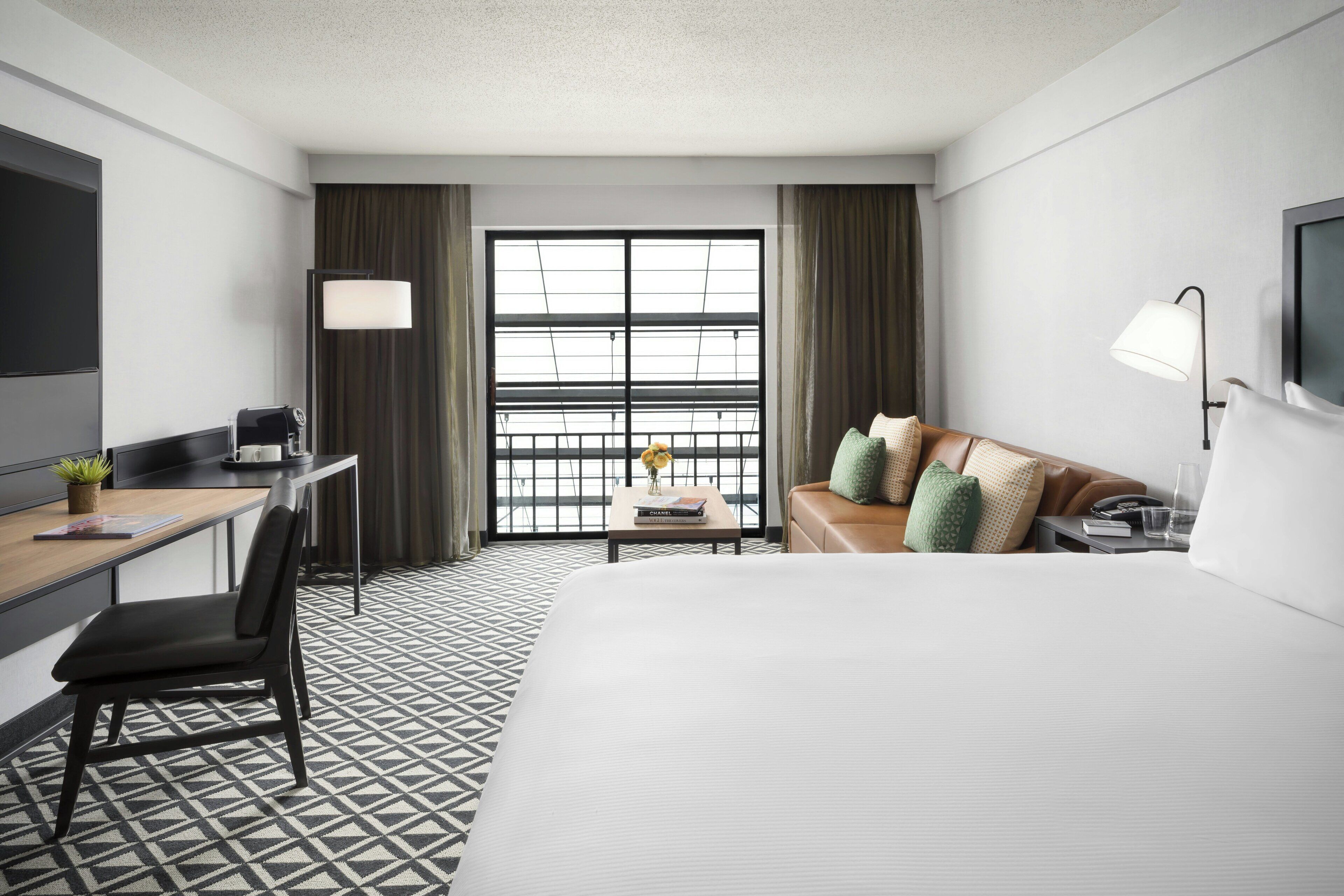 Ein Hotelzimmer mit einem Bett, einem Schreibtisch mit Stuhl, einer Couch mit Kissen, einem Fernseher, einem Fenster mit Vorhängen und einem gemusterten Boden.