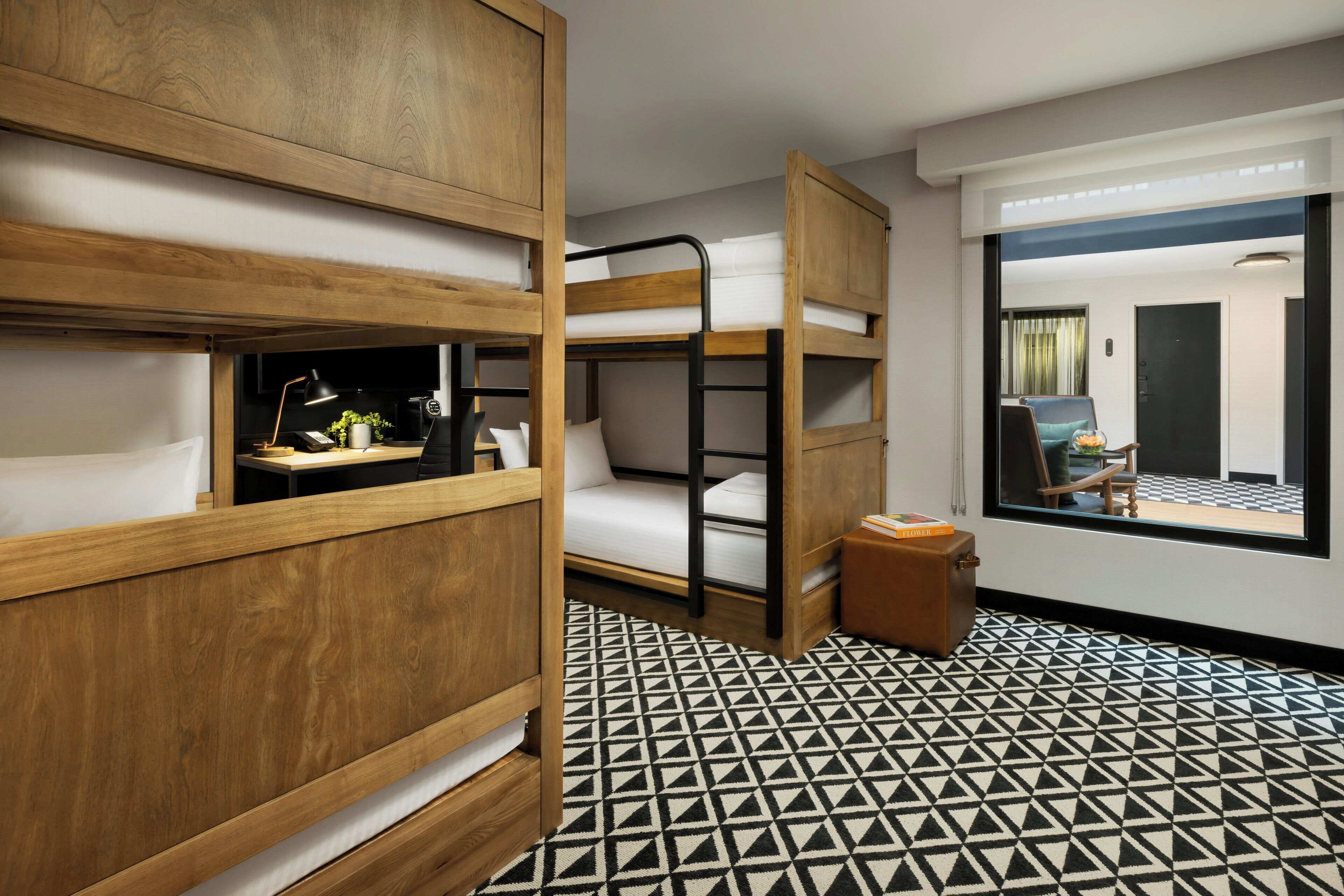 A hotel room with bunk beds, a desk with a lamp, a small table with a plant, and a window with a view of the outside.