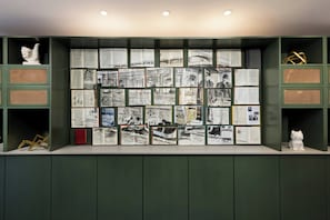 A wall with multiple framed photographs and documents, a white sculpture, and a red book on a shelf. - Arcadian Hotel (Brookline)