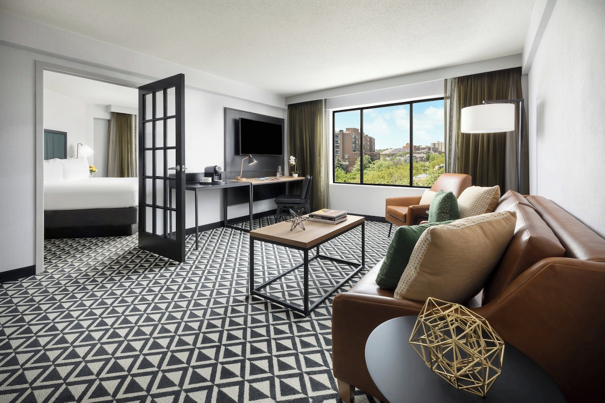 A hotel room with a bed, a desk, a sofa, and a large window offering a city view.