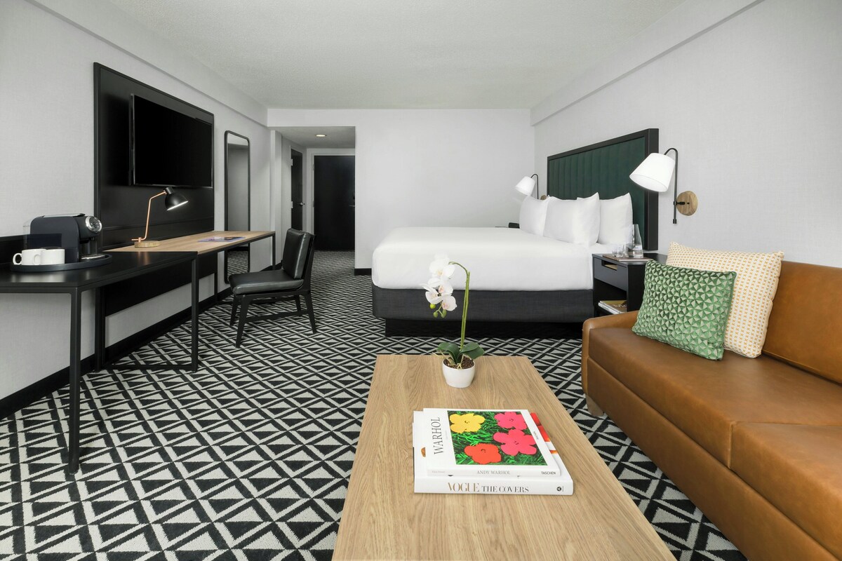 A hotel room with a bed, a desk with a coffee machine, a TV, a sofa, and a coffee table with a magazine.