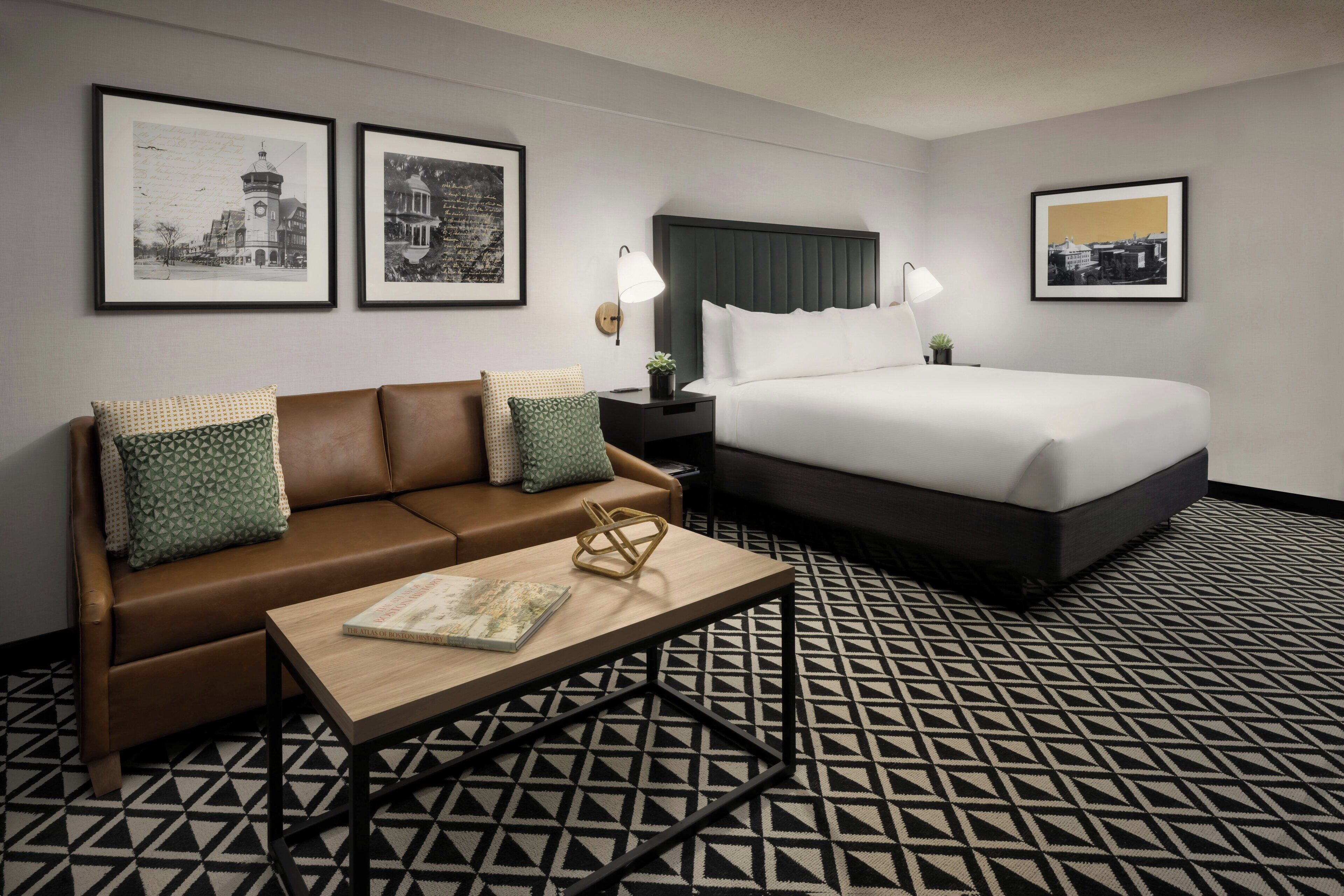 Habitación de hotel con un sofá de cuero marrón, una mesa de centro de madera con un libro, una cama con ropa de cama blanca y dos fotografías blanco y negro enmarcadas en la pared.