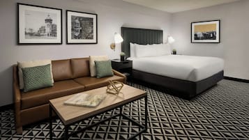 Habitación de hotel con un sofá de cuero marrón, una mesa de centro de madera con un libro, una cama con ropa de cama blanca y dos fotografías blanco y negro enmarcadas en la pared.