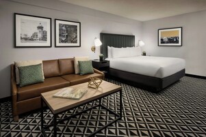 Habitación de hotel con un sofá de cuero marrón, una mesa de centro de madera con un libro, una cama con ropa de cama blanca y dos fotografías blanco y negro enmarcadas en la pared.