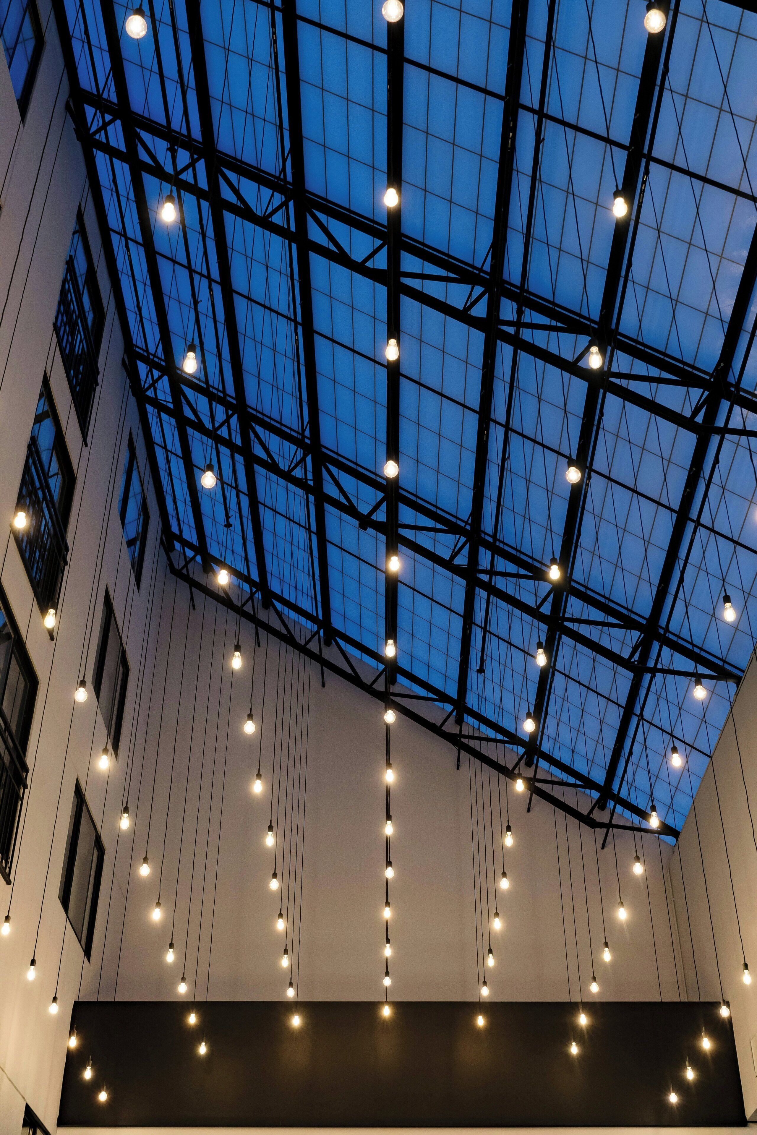 A modern building interior with a high ceiling, exposed structural beams, and numerous hanging light fixtures.