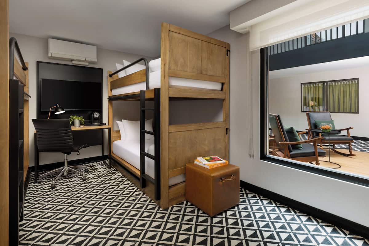 A compact hotel room with a bunk bed, a desk with a chair, a small wooden cabinet, and a large window offering a view of the outside.