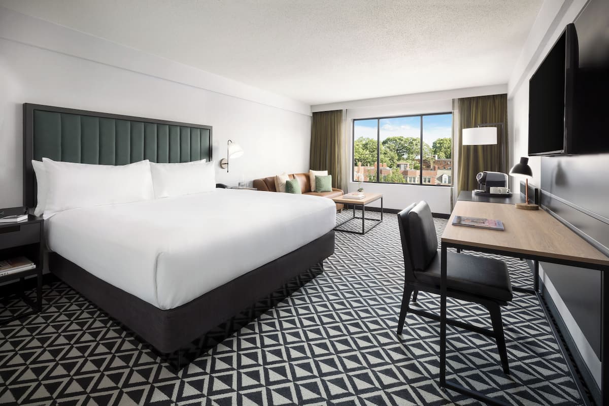 A hotel room with a large bed, a desk with a chair, a TV, and a window with a view of buildings.
