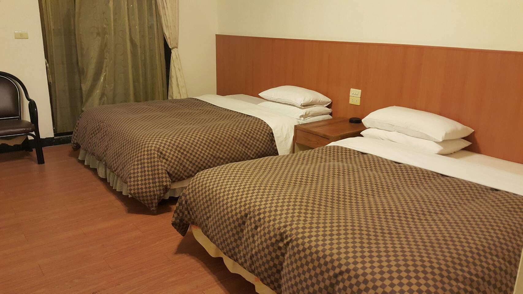 A hotel room with two single beds, wooden headboards, a wooden nightstand, a chair, and a curtain.