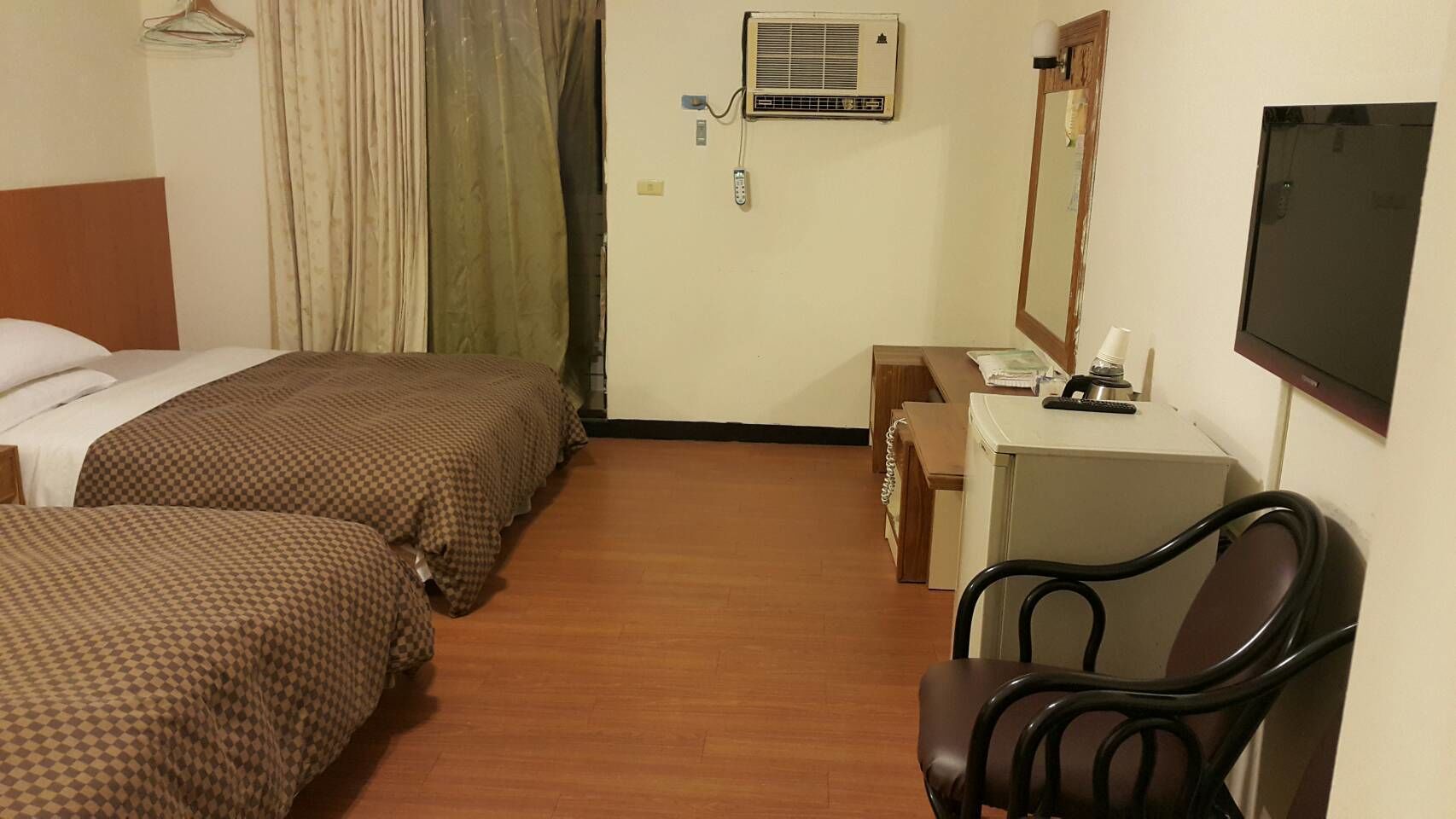 A hotel room with two beds, a desk with a chair, a mirror, an air conditioning unit, and a television.