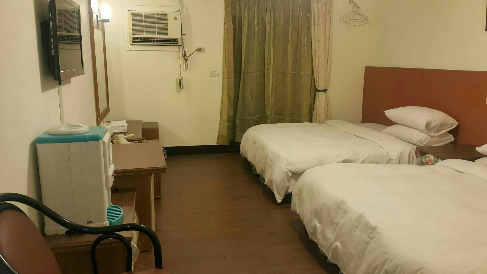 Habitación de hotel con dos camas, un escritorio con silla, un calentador y una ventana con cortinas.