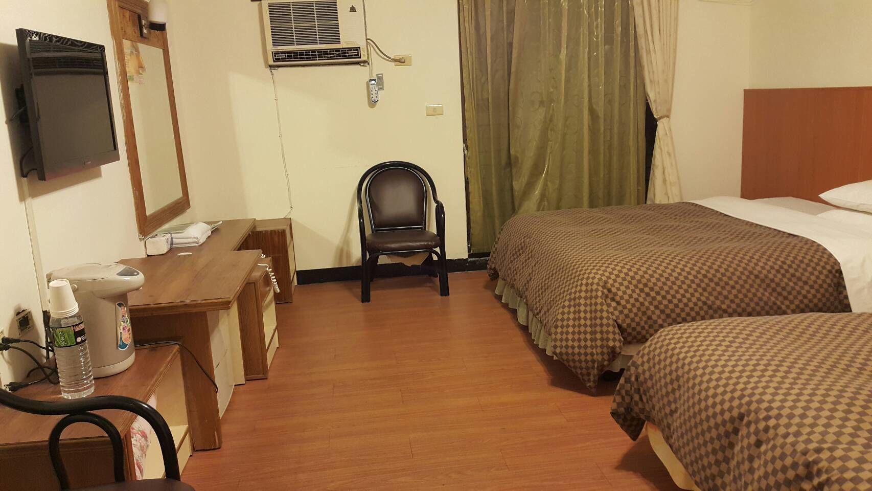 A hotel room with a desk, chair, two beds, a television, and an air conditioning unit.