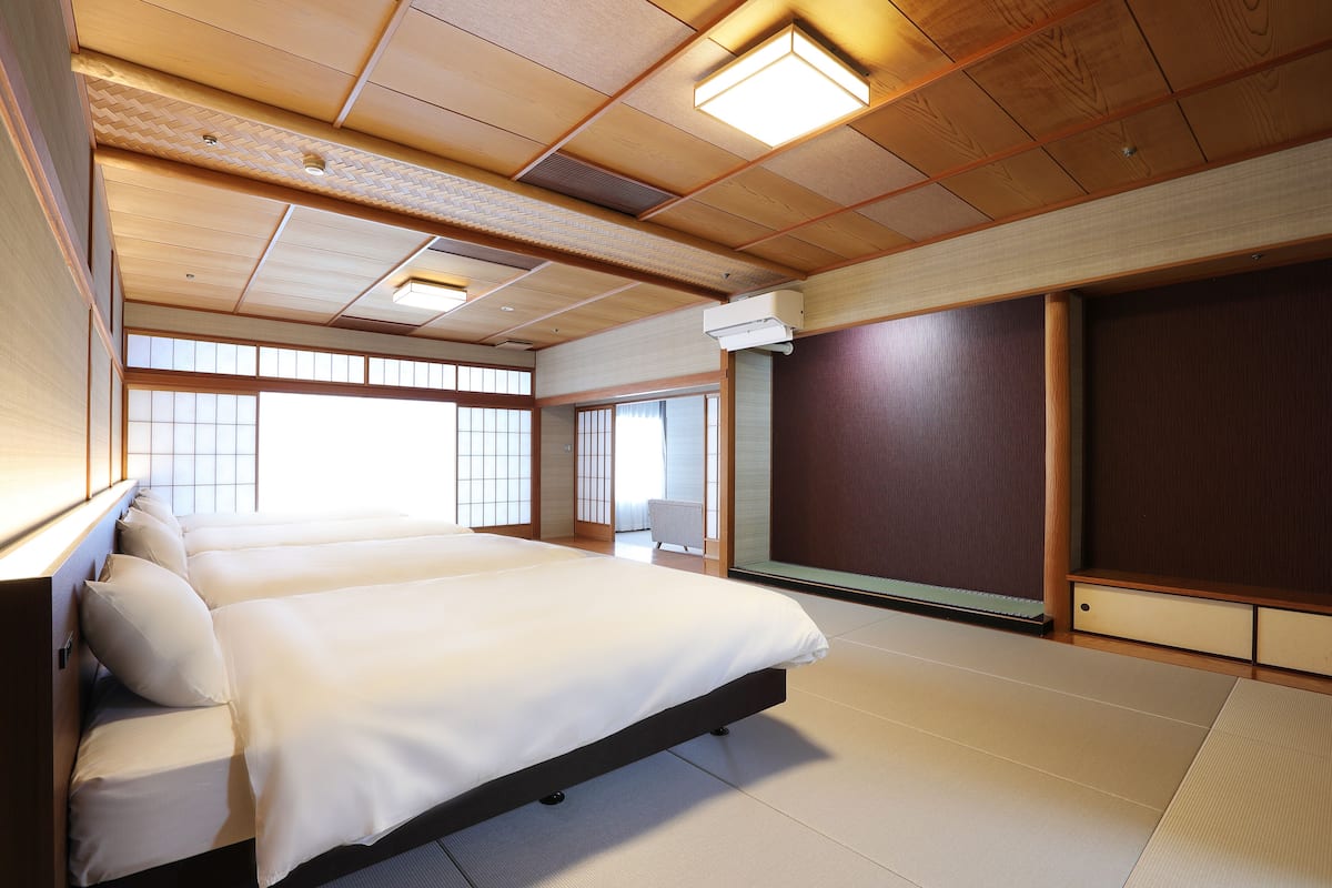 A traditional Japanese-style bedroom with tatami flooring, a large bed with white bedding, sliding doors, and a ceiling with wooden beams and recessed lighting.