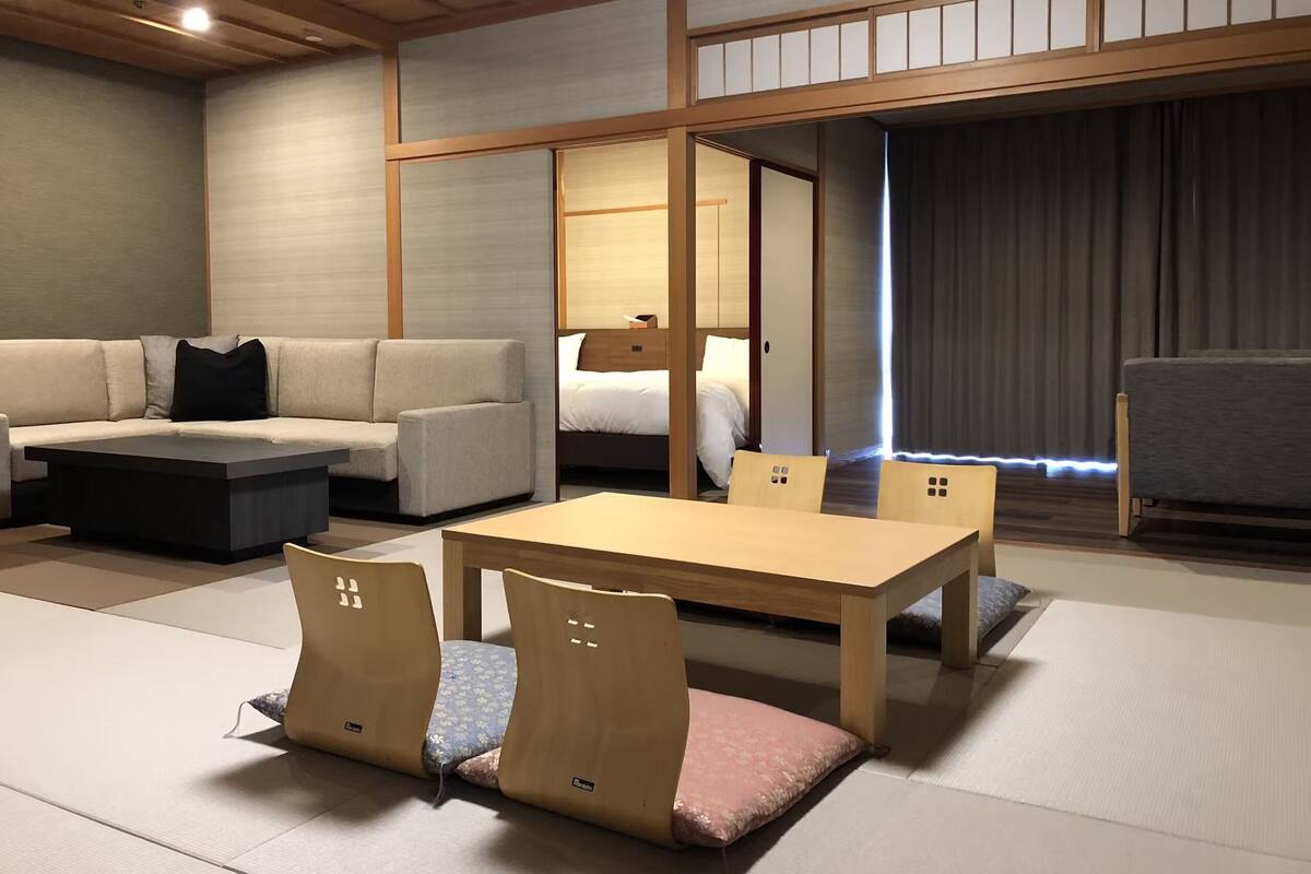 A traditional Japanese-style room with tatami flooring, a low wooden table with chairs, a sofa, and a bed with a wooden headboard.