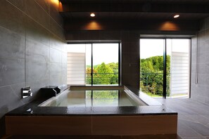 A modern bathroom with a glass-enclosed bathtub, large windows, and a view of greenery outside. - Manten no Tsujinoya (Komatsu)