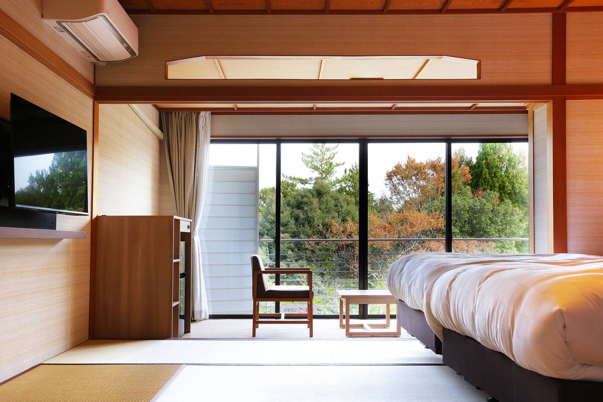 A modern bedroom with a bed, a chair, a small table, a TV, and a large window with a view of trees.