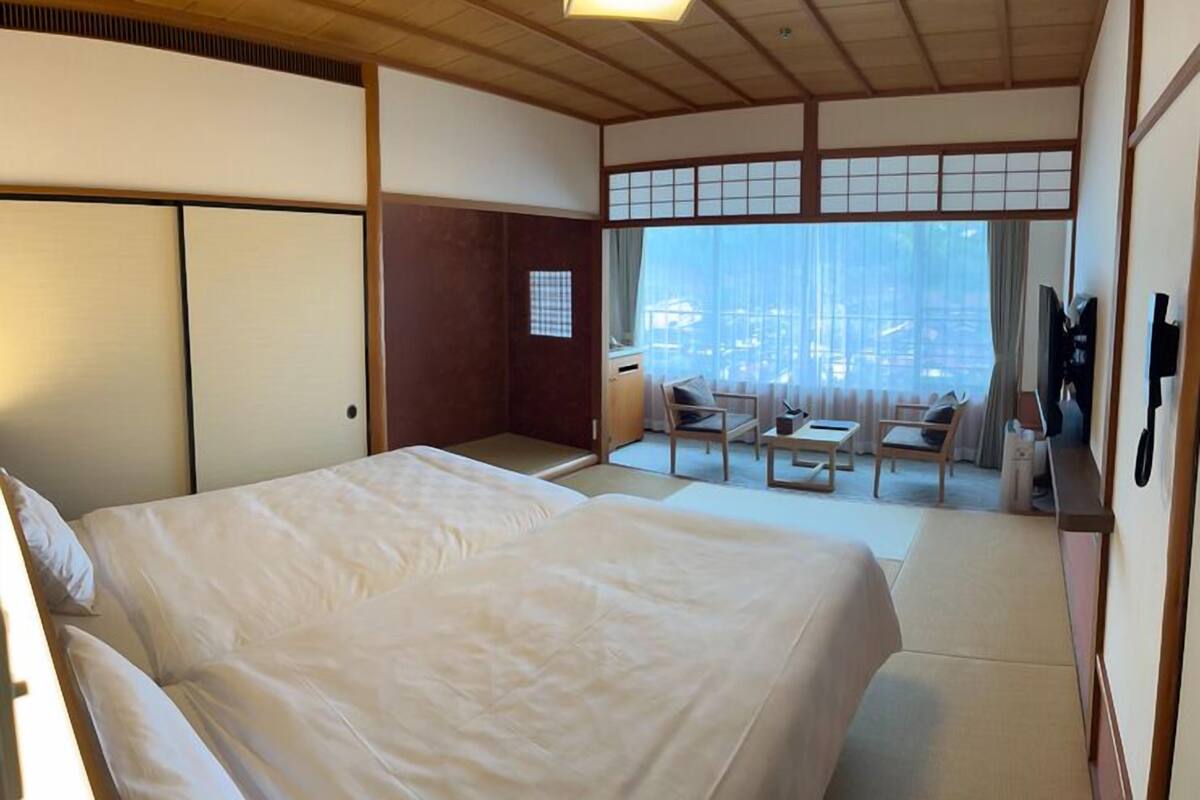 A hotel room with a large bed, a small table, a chair, and a view of the city through large windows.