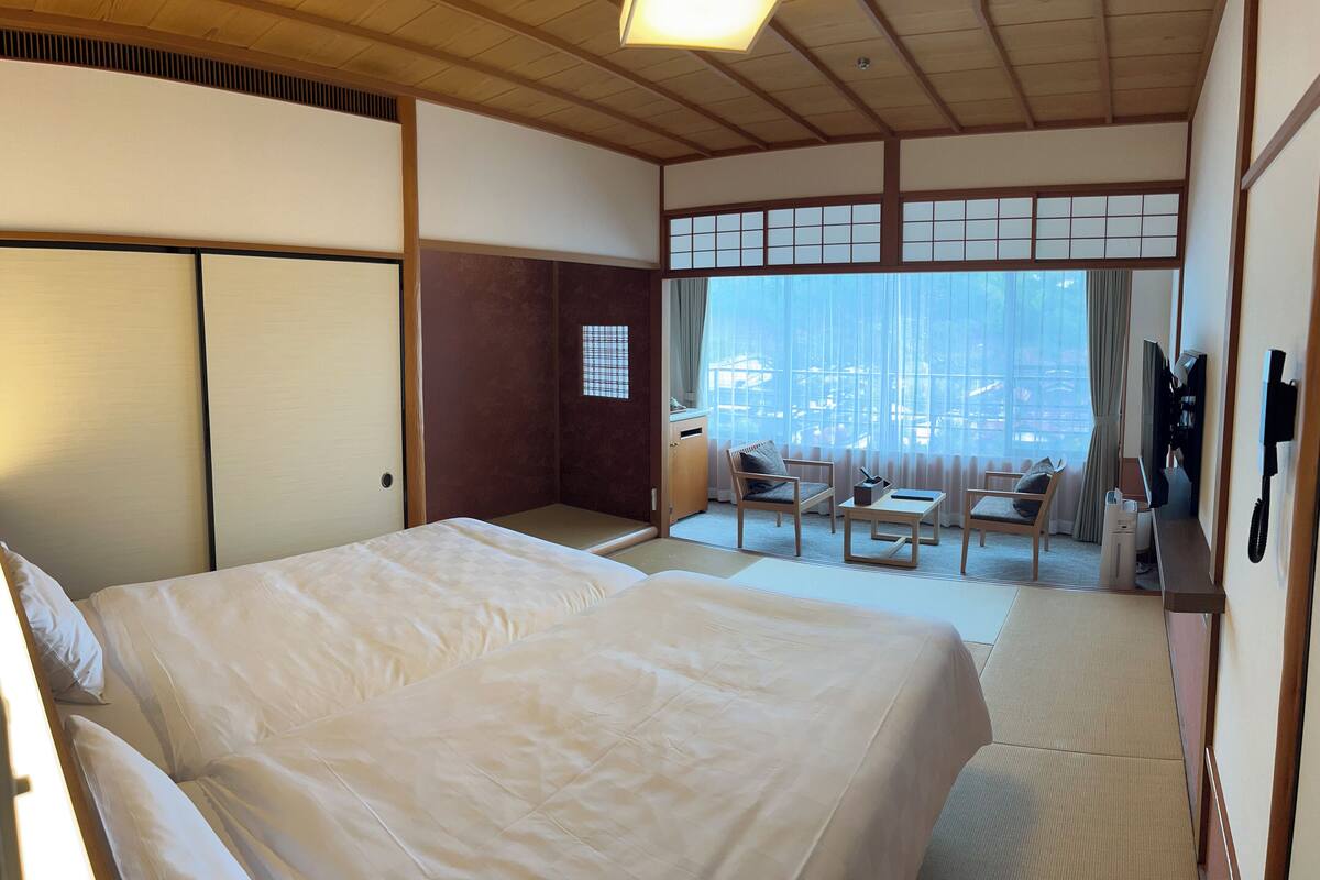 A traditional Japanese-style hotel room with tatami flooring, a large bed, a small table with chairs, and a view of the city through large windows.