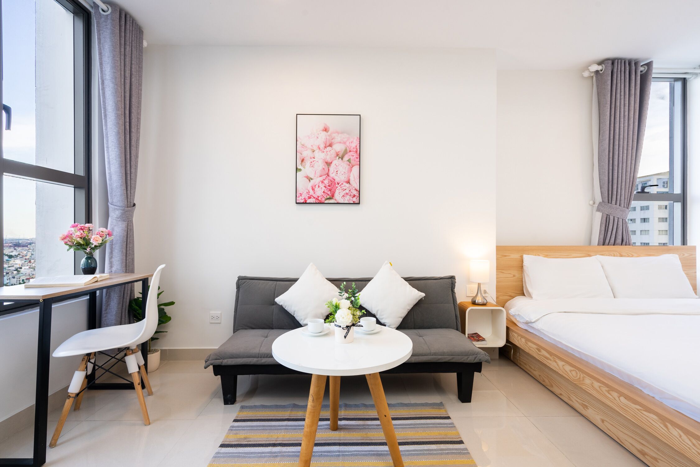 A modern hotel room with a bed, a sofa, a dining table, and a chair. There is a window with curtains, a painting on the wall, and a small table with a vase of flowers.