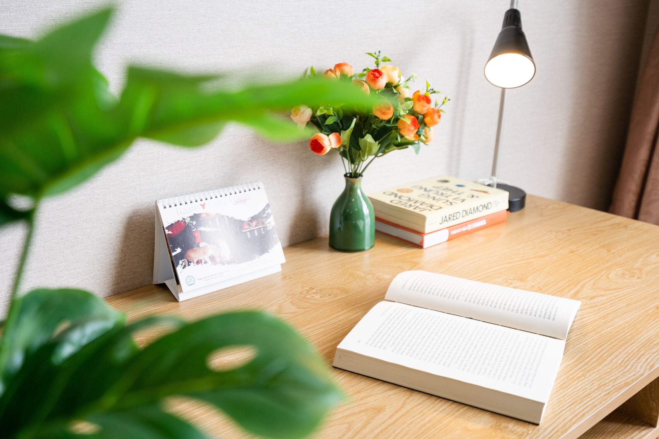 A wooden table with a book, a vase with flowers, a calendar, and a lamp.
