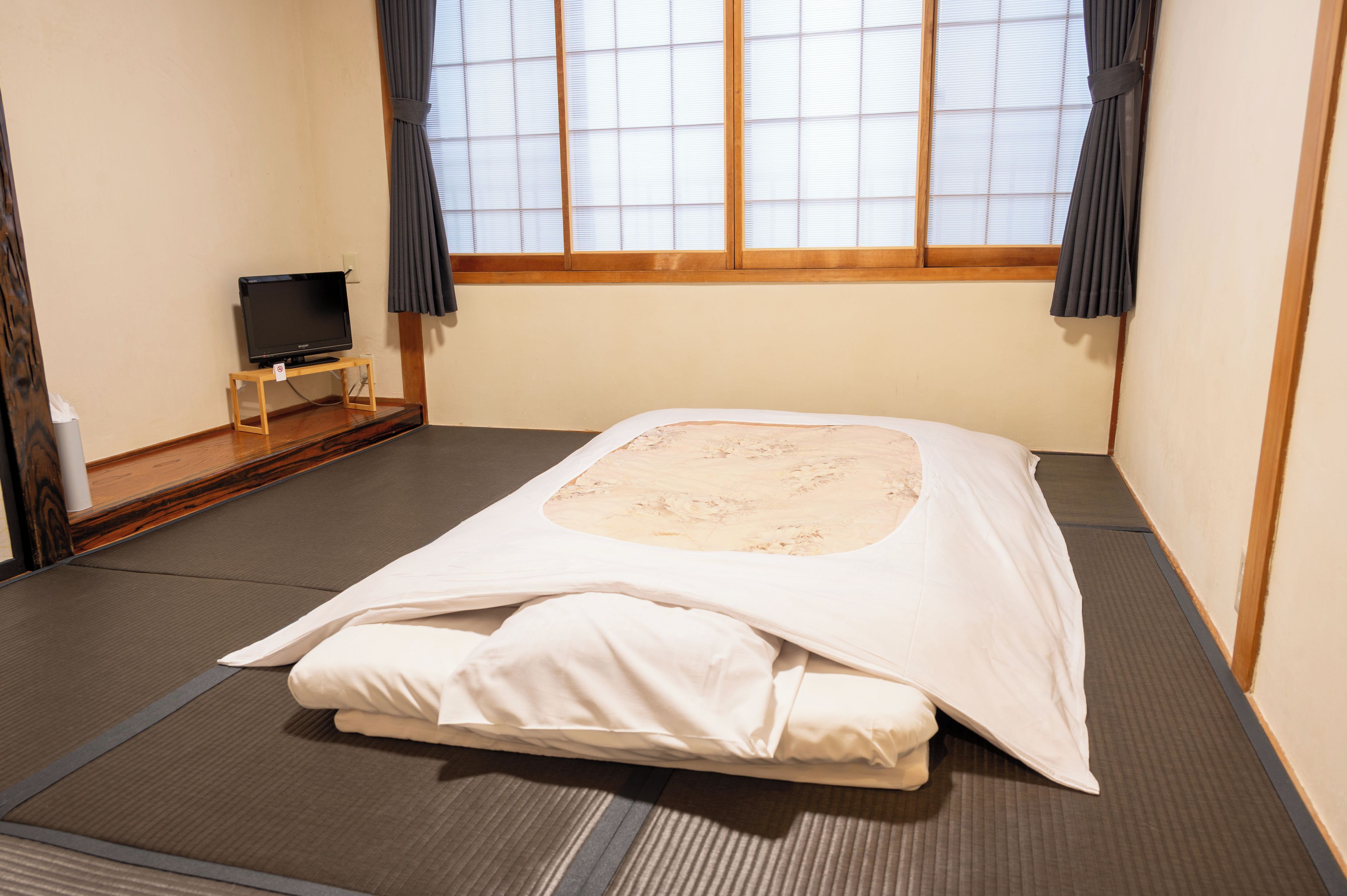 Una habitación con suelo de tatami, una cama con ropa blanca, un televisor sobre un pequeño soporte y una ventana con persianas.