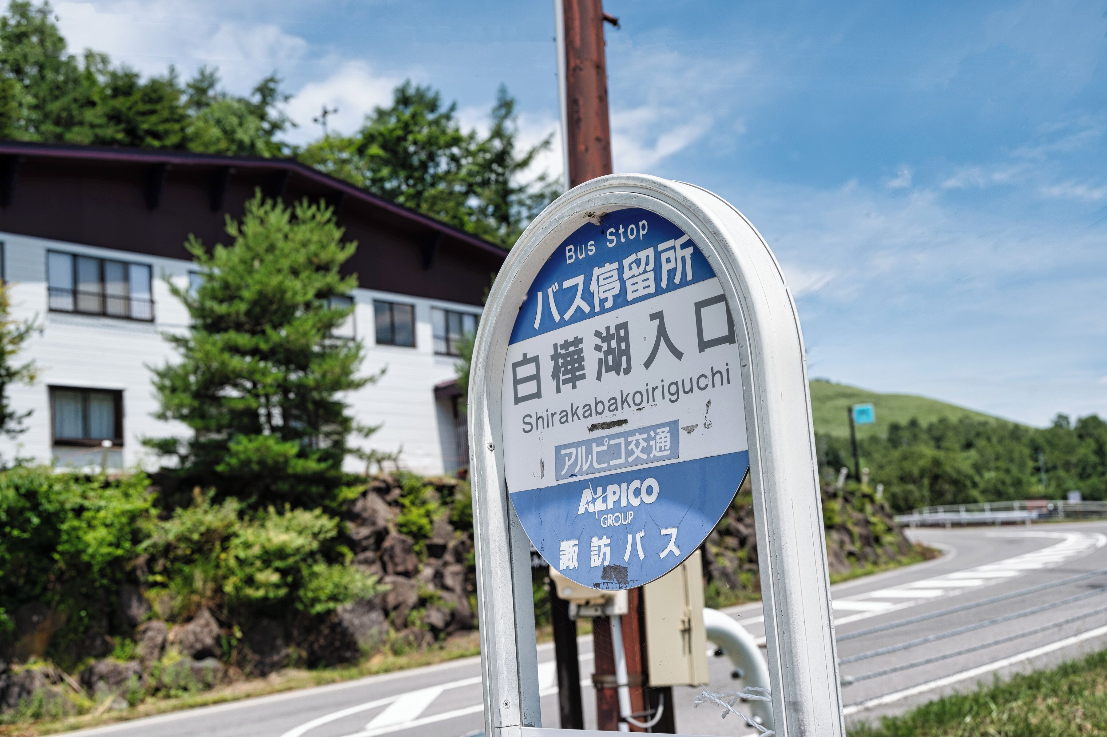 Una señal de parada de autobús con texto en japonés, un edificio de techo rojo y una carretera con una curva.