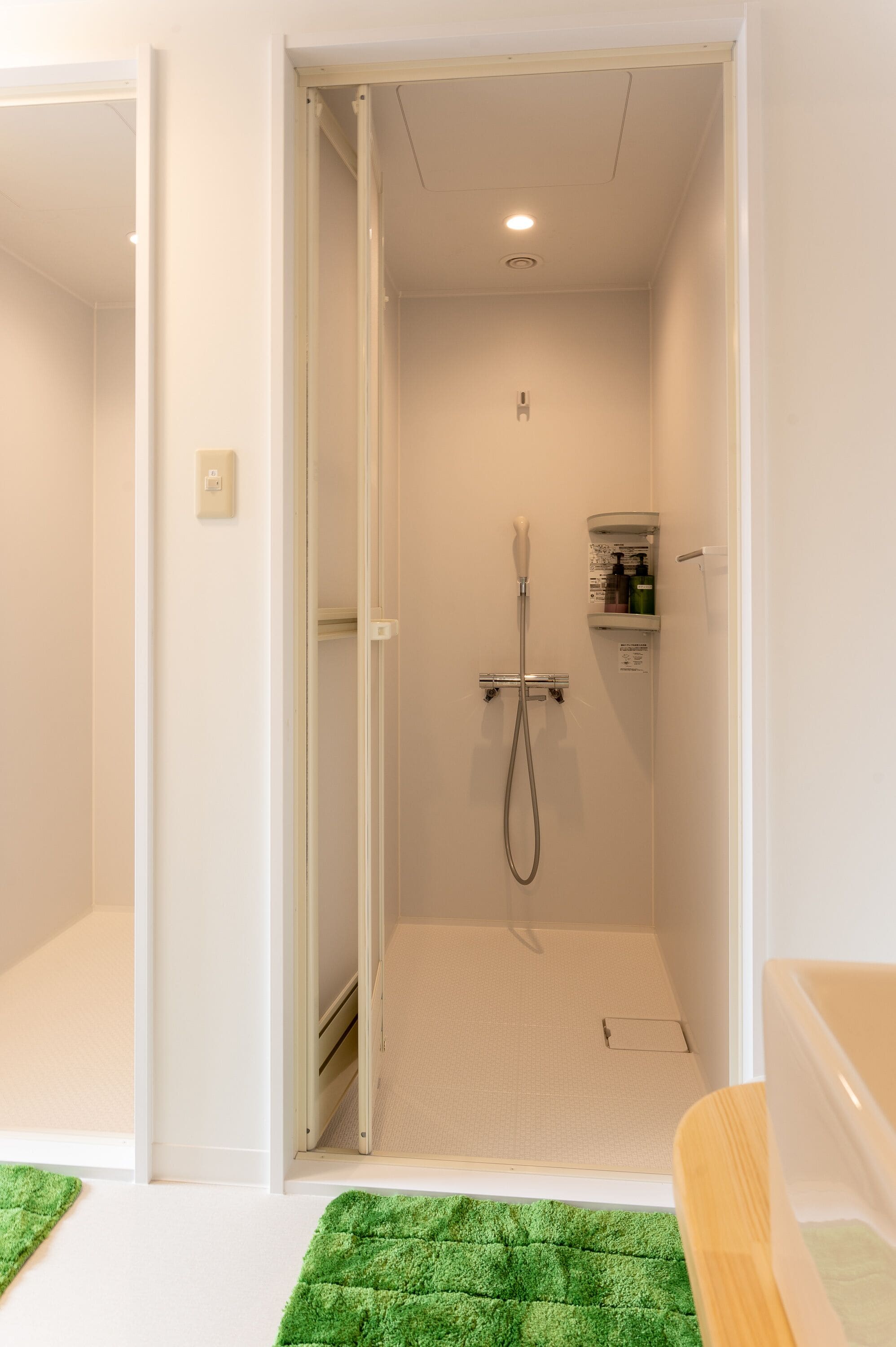 Una doccia a pavimento con soffione estraibile, un piccolo ripiano con articoli da toilette e un tappeto verde sul pavimento.