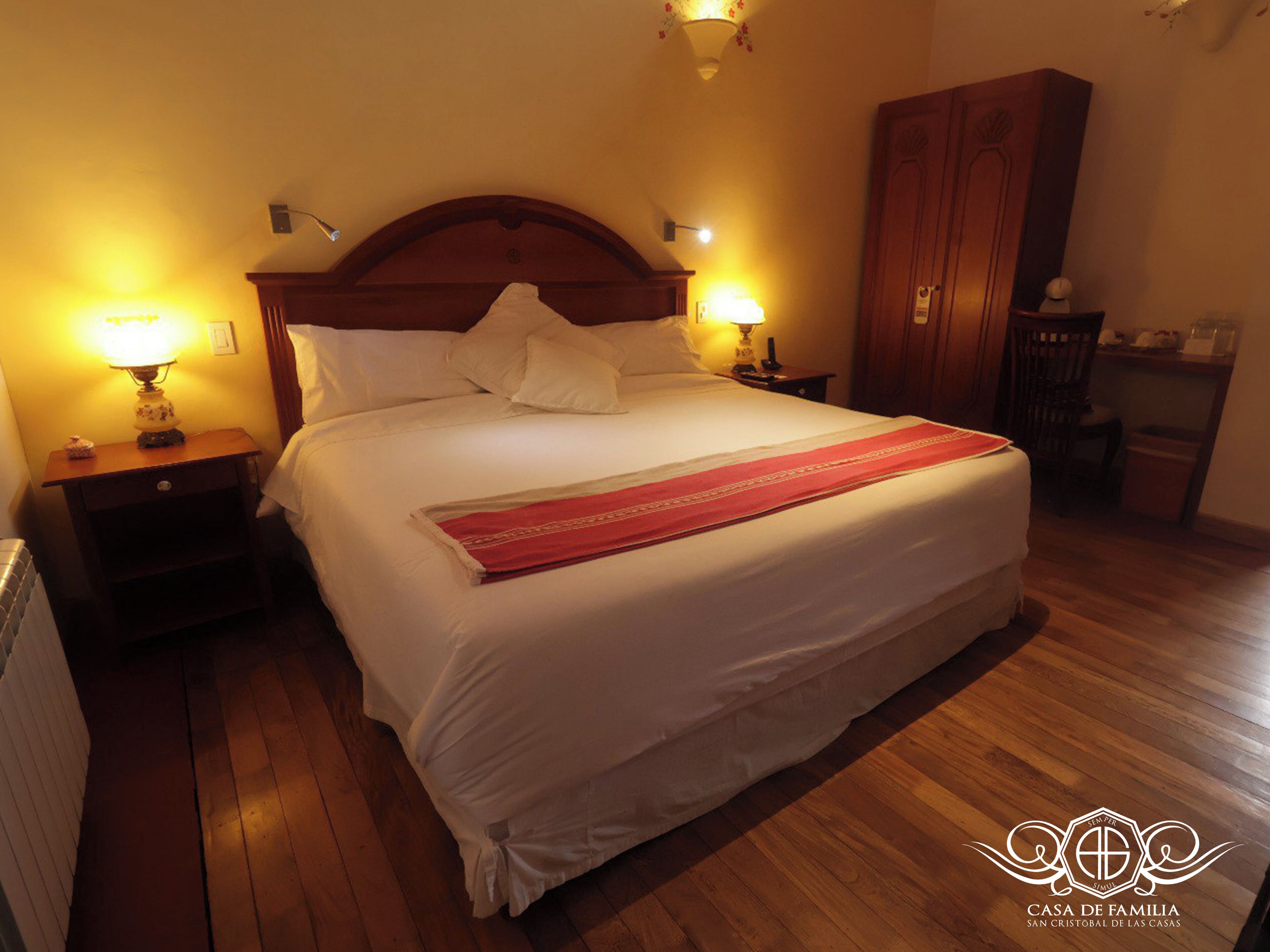 A neatly made bed with white linens and a red runner, flanked by two bedside lamps.