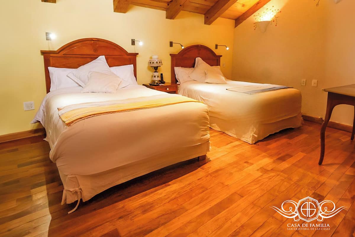 Quarto com duas camas, piso de madeira e mesa de cabeceira com abajur.