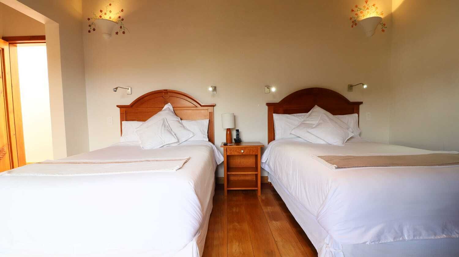 A hotel room with two single beds, wooden headboards, bedside tables with lamps, and a small wooden nightstand.